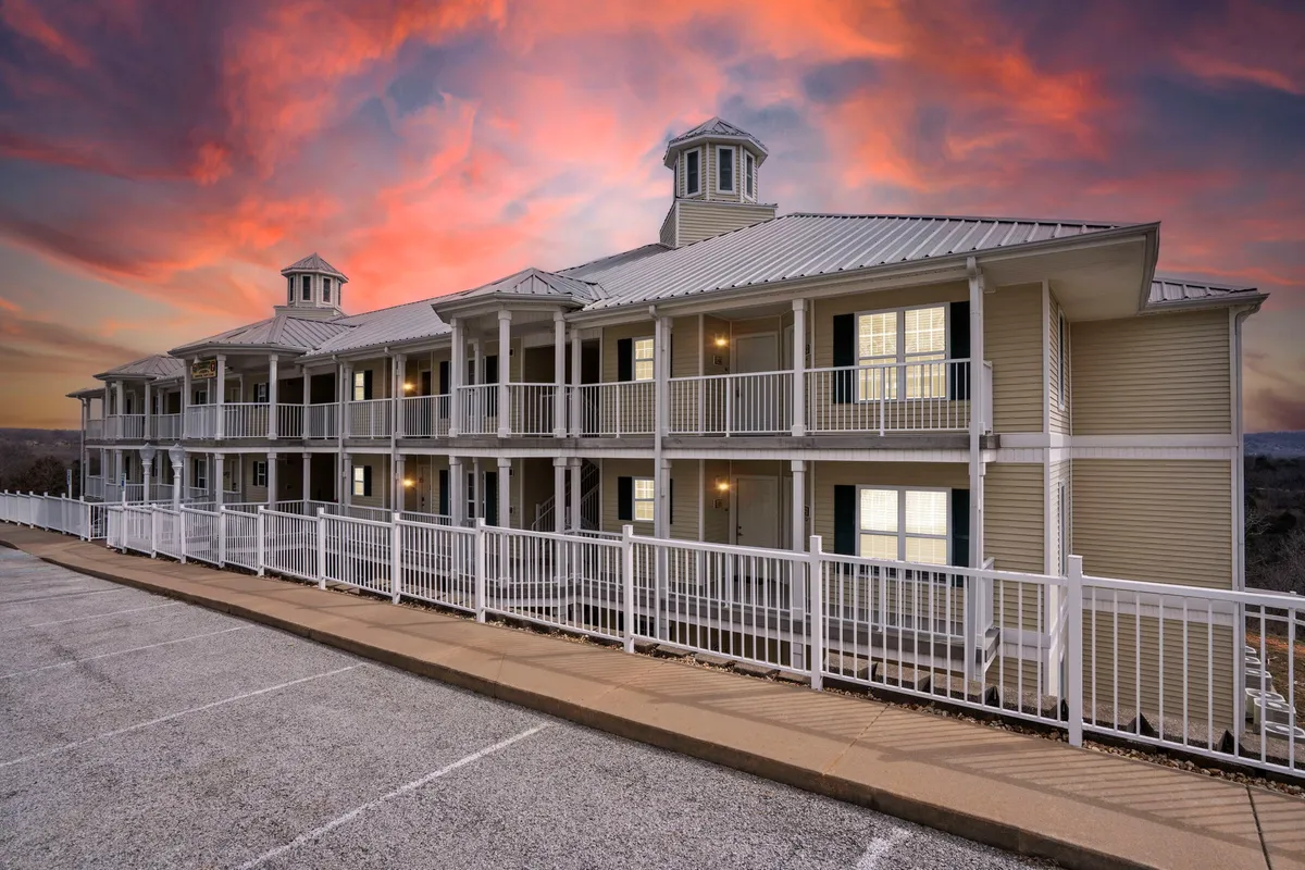 This image features a multi-story resort building with detailed architecture, set against a colorful sunset sky. People are seen strolling along the well-lit pathways, enjoying the ambiance, and socializing on balconies, which overlook the tranquil resort grounds.