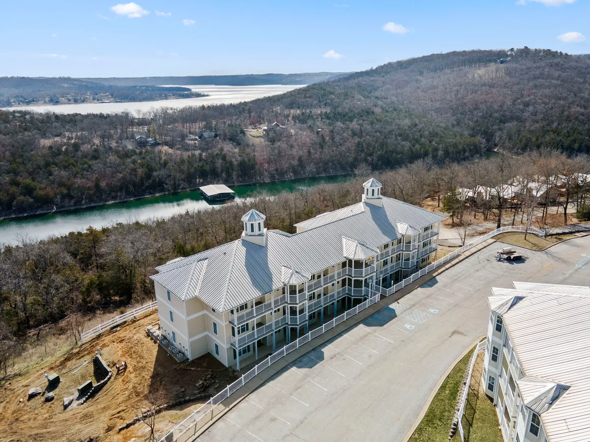 An exterior view captures the property’s striking architecture set beside a tranquil body of water and surrounded by rolling hills. The image evokes the natural beauty of the Ozarks and the opportunities for hiking, boating, and enjoying the area's vibrant fall foliage.