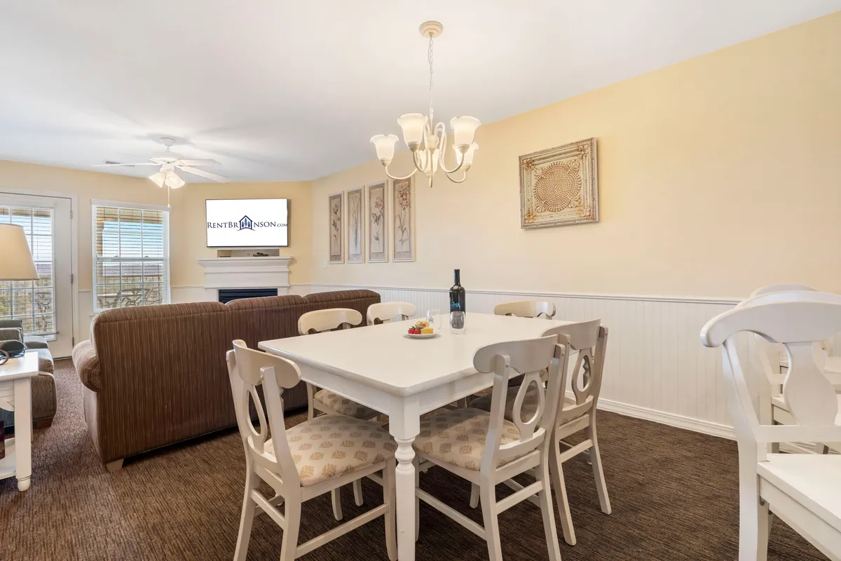 A warm and inviting living/dining area featuring a well-set dining table, a cozy sofa, a chandelier overhead, and wall-mounted television, all accentuated by homey artwork. The space is open and designed for gatherings and relaxation, reflecting the comfort and newness of the home.
