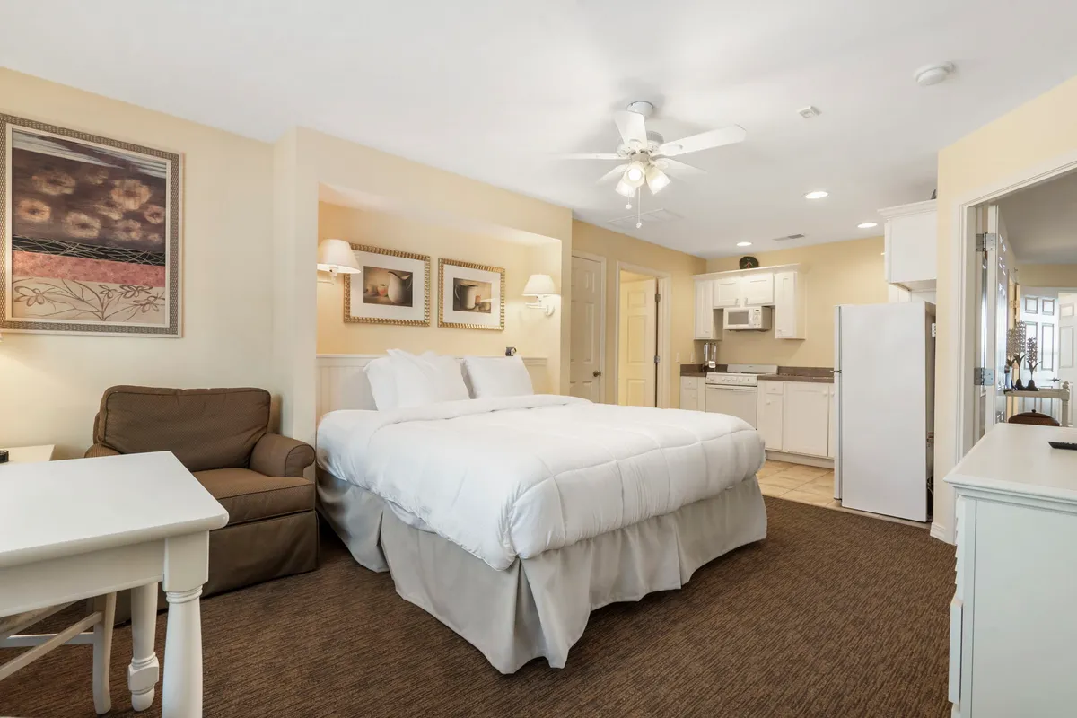 A king bedroom with a large, neatly made bed and crisp white bedding, complemented by a comfortable armchair, a small kitchenette area, and thoughtfully chosen artwork on the walls. The space feels calm, organized, and homelike.
