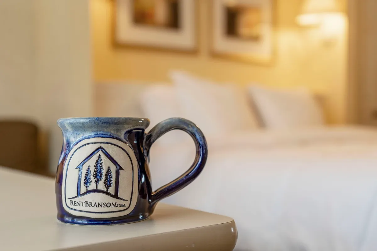A close-up of a cozy bedroom scene with a ceramic coffee mug on the bedside table, crisp white linens, and framed pictures on the wall. Soft natural light enhances the ambiance, offering a sense of peace and relaxation.