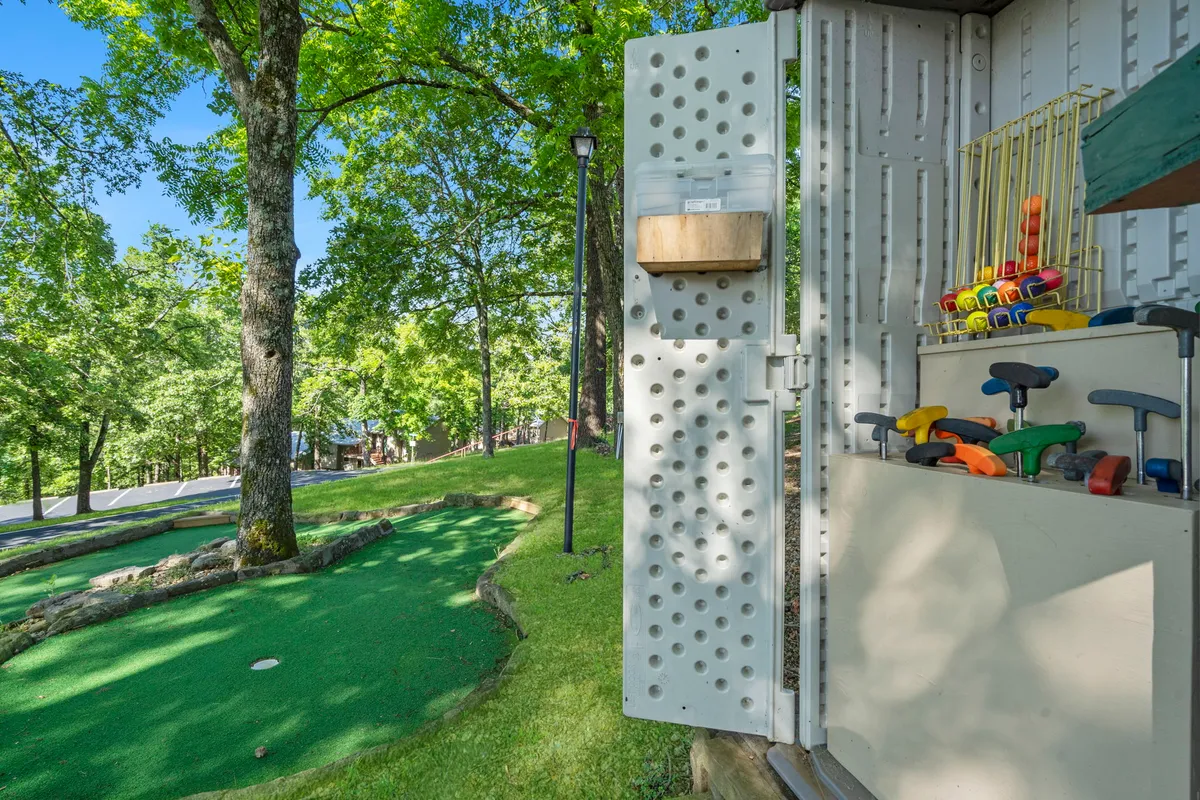 A playful mini-golf putting area set amidst green grass and trees, with families enjoying friendly competition and laughter. A storage shed adds a touch of resort practicality.
