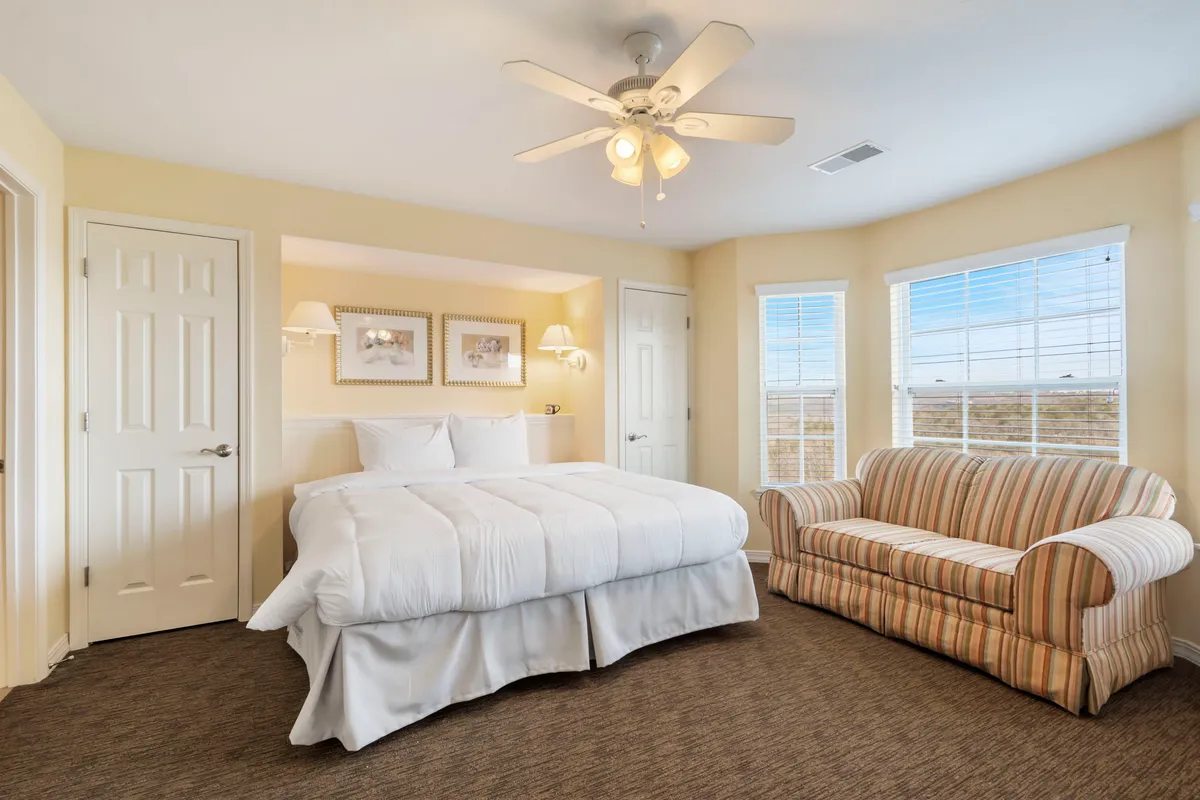 A spacious bedroom featuring a large, inviting bed with plush white bedding, a striped couch by the sunlit window, and soft natural light streaming in. The calming decor and cozy furniture create a tranquil atmosphere, perfect for rest and relaxation.