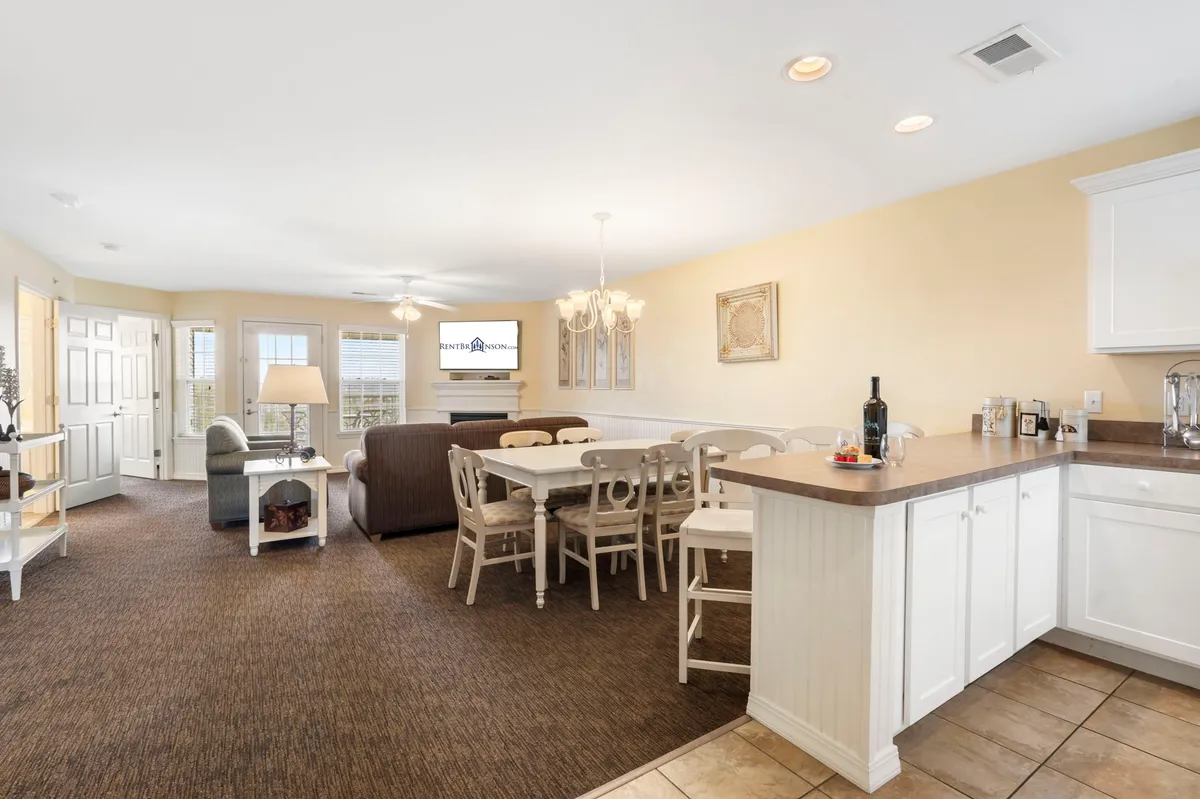 An inviting open-concept room that seamlessly blends the living area, dining table, and kitchen counter. The seating area with a sofa is adjacent to both the dining table and a visible TV, with a chandelier hanging above the dining space, creating a sense of warmth and togetherness.