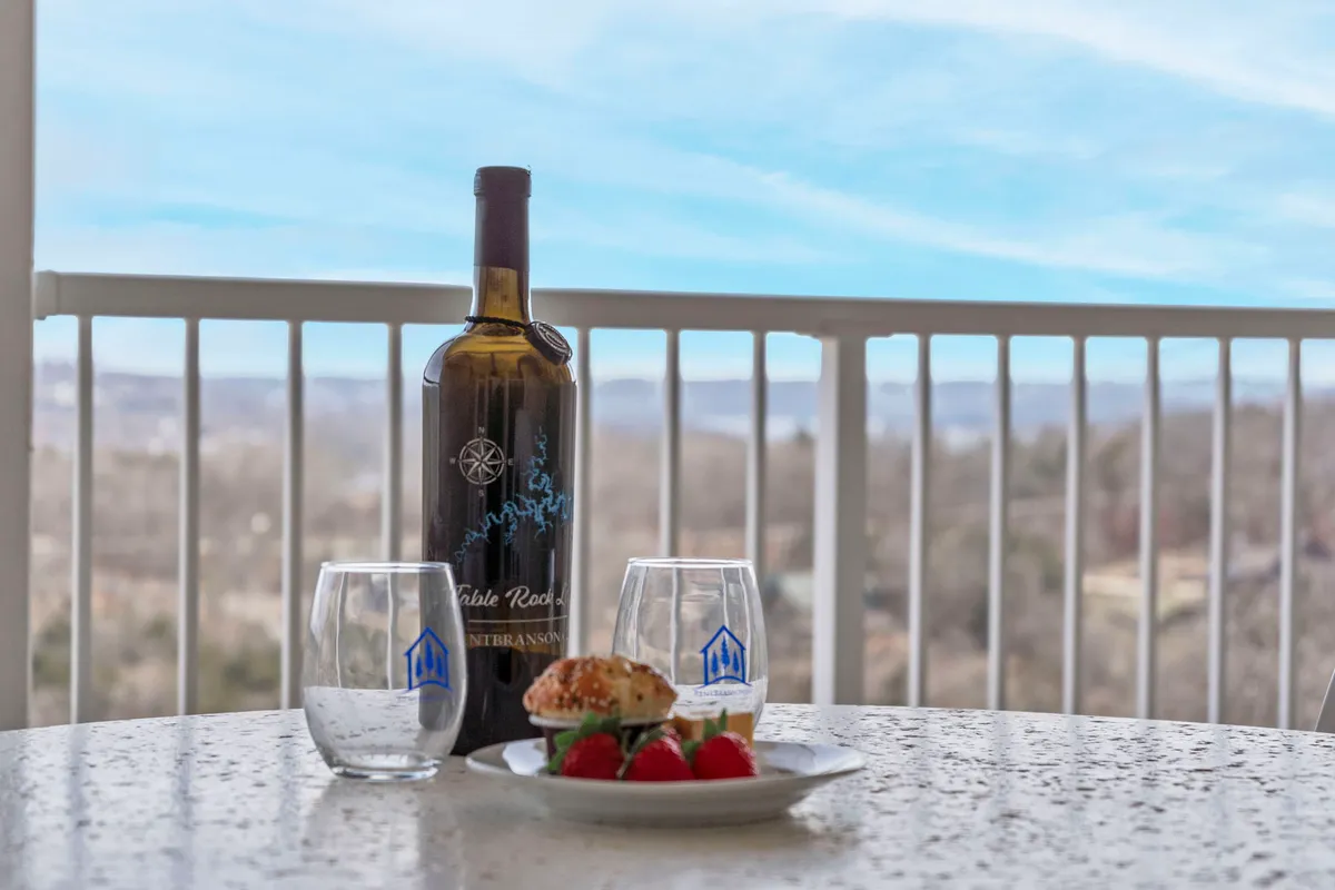 A close-up view of a wine bottle, glasses, and a plate with strawberries and a scone, arranged on a table with a scenic backdrop. The composition evokes a sense of quiet relaxation and indulgence, perfect for a romantic setting.