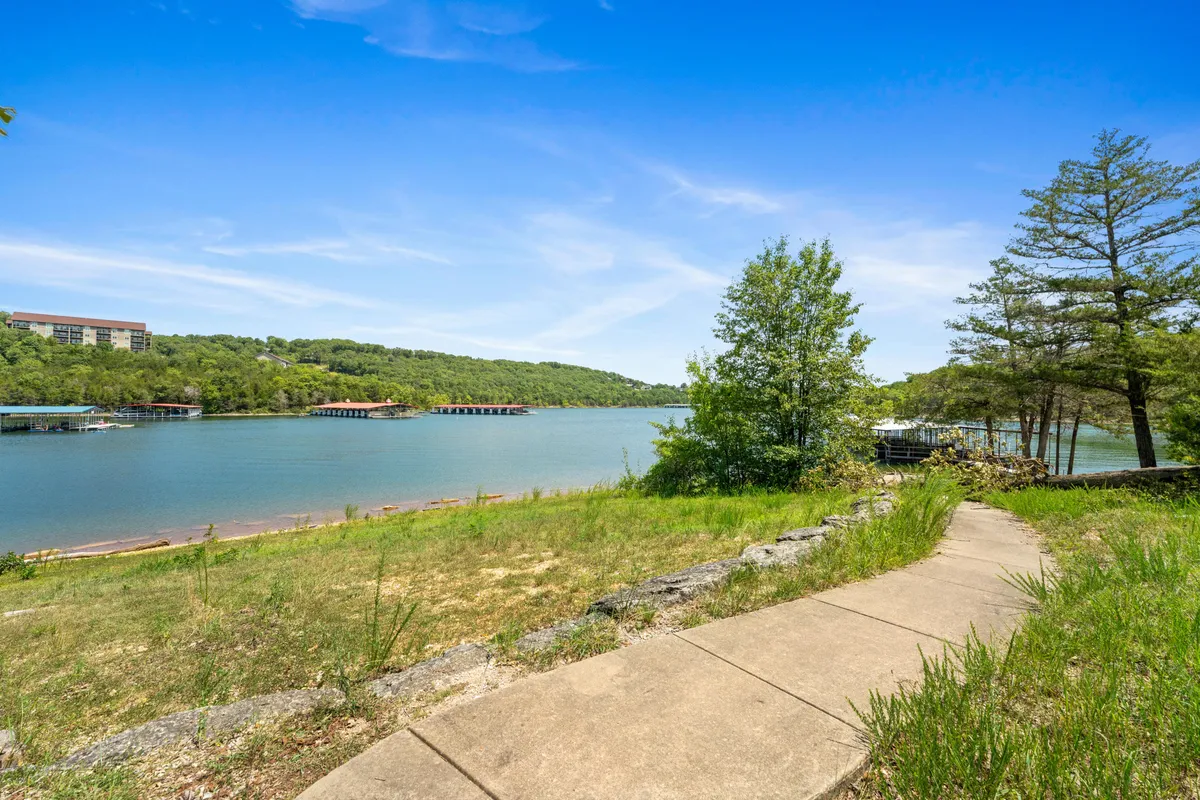 A tranquil exterior pathway winds along the lake, bordered by trees and gentle hills in the background. The image conveys natural beauty and easy access to walking trails, ideal for strolls with pets or family members.