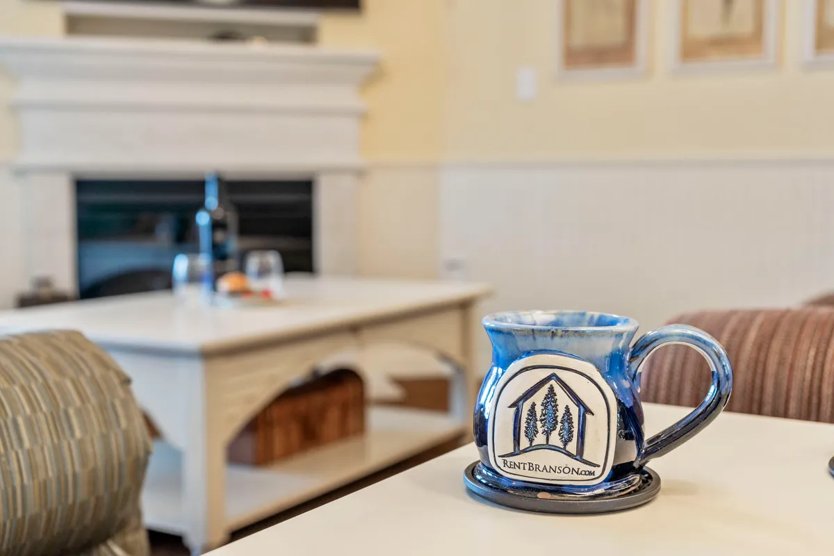 A cozy interior scene centers on a ceramic mug resting on a coffee table in front of a glowing electric fireplace. The setting radiates comfort and warmth, ideal for cooler seasons, and hints at intimate moments of relaxation, reading, or sharing stories with friends.